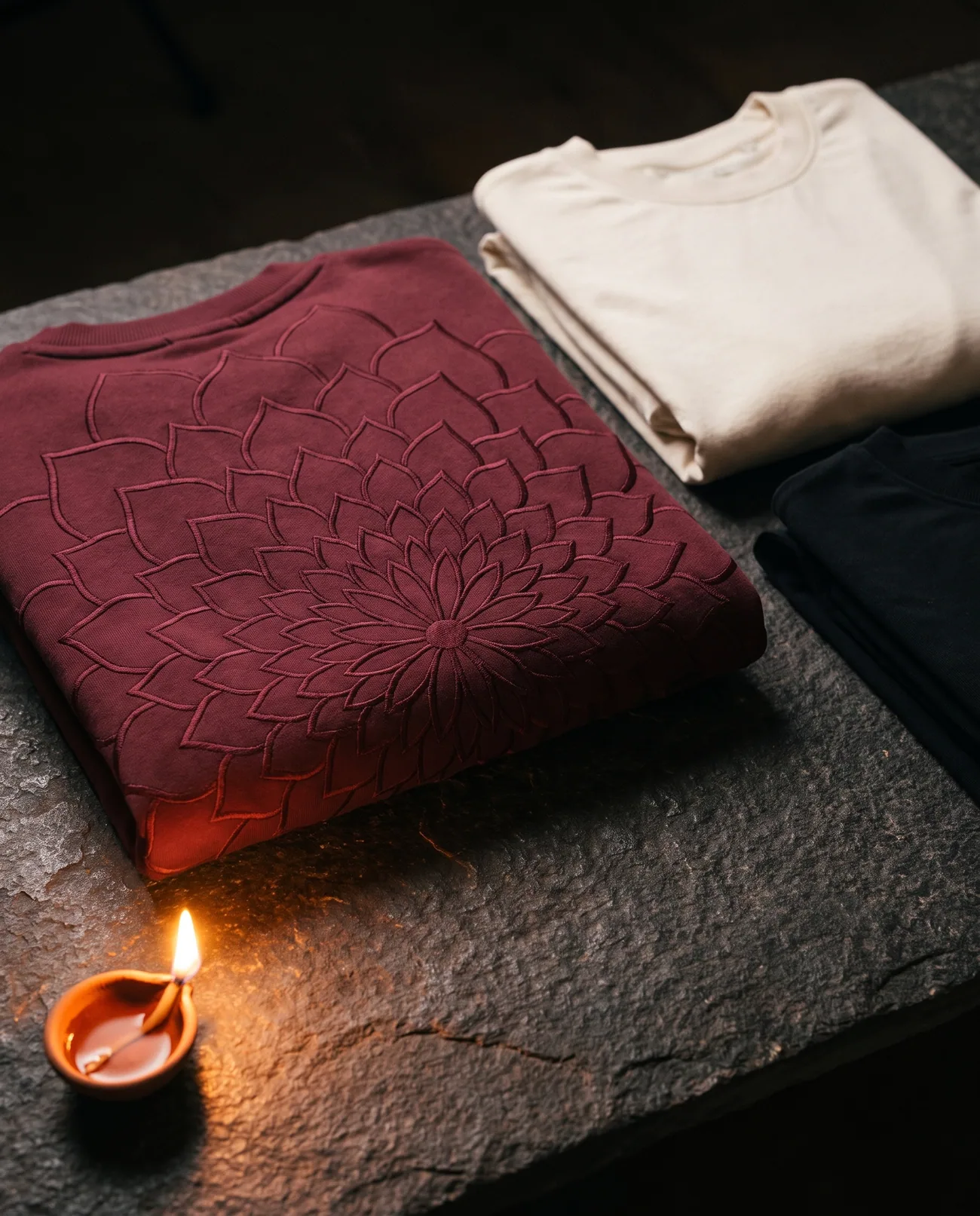 The Rangoli Sweatshirt folded on dark stone beside a lit diya
